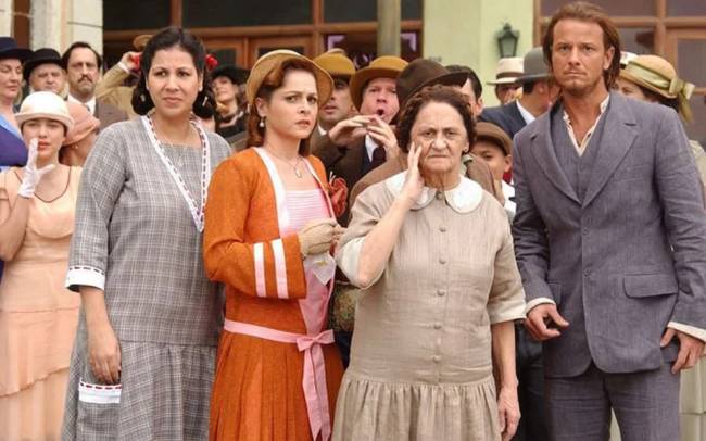 Grupo de pessoas com roupas de &eacute;poca, incluindo tr&ecirc;s mulheres e um homem em primeiro plano, com express&otilde;es s&eacute;rias e preocupadas. A mulher &agrave; esquerda veste um vestido xadrez cinza, a do meio um casaco laranja sobre um vestido rosa, e a senhora mais velha, &agrave; direita, um vestido bege com gola branca, tocando o rosto. O homem &agrave; direita veste um terno cinza.