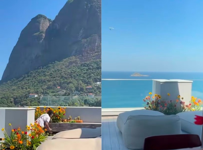 Homem de camisa branca agacha em terraço com vista para montanha verde e mar azul, com ilha ao fundo. Há vasos de flores coloridas e pufes brancos no ambiente externo