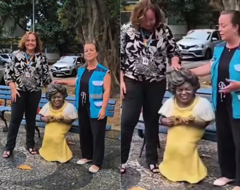 Duas mulheres sorridentes, uma de blusa estampada e outra de colete azul, posam ao lado de uma est&aacute;tua de mulher negra sentada em um banco, vestindo um vestido amarelo e segurando um objeto. A mulher de blusa estampada toca a cabe&ccedil;a da est&aacute;tua, enquanto a de colete azul gesticula. O cen&aacute;rio &eacute; uma cal&ccedil;ada arborizada com carros ao fundo
