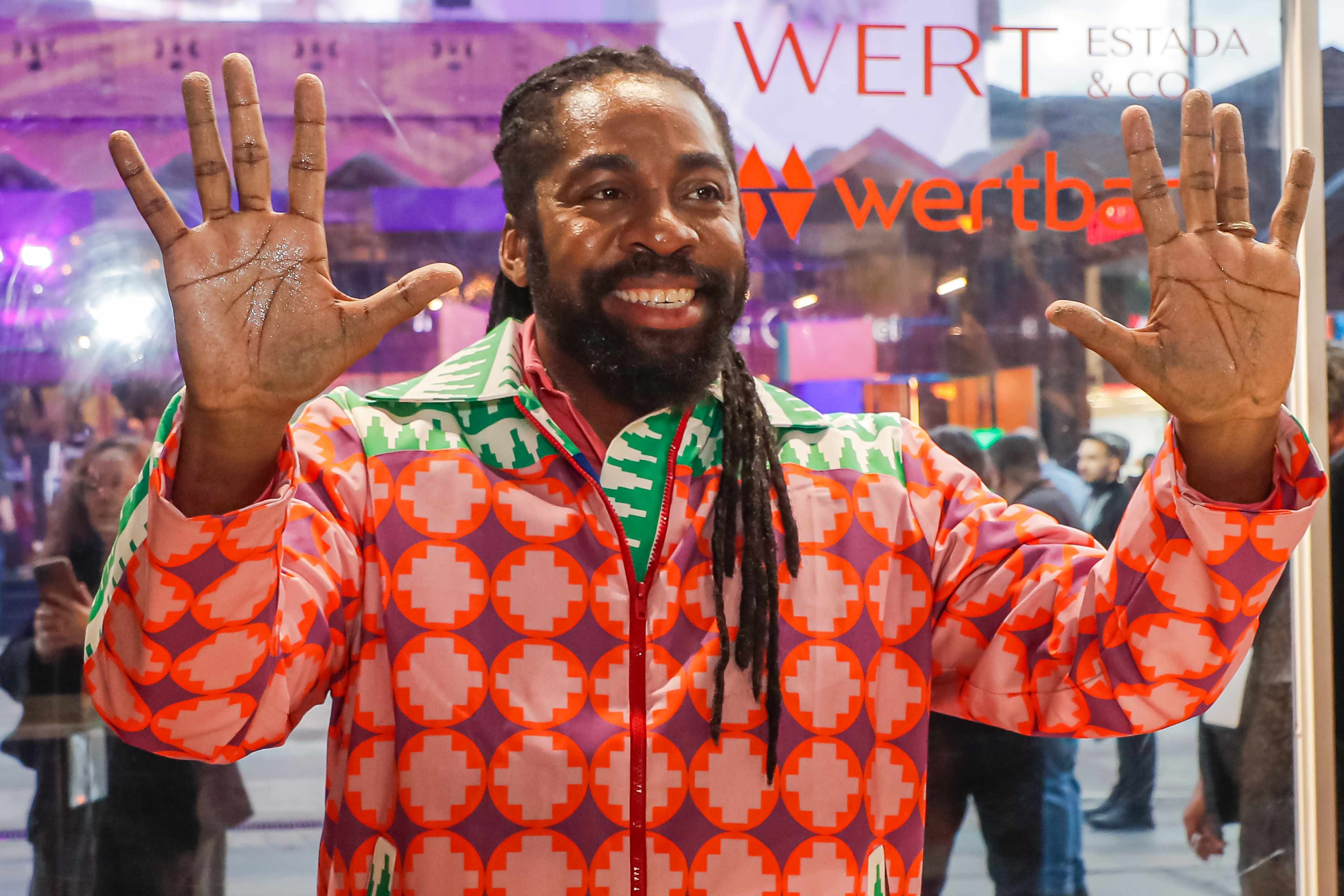 Homem negro de barba e dreadlocks sorri, vestindo jaqueta colorida com estampa geométrica laranja, rosa e roxa, levantando as mãos abertas com glitter nas palmas. Ao fundo, uma vitrine reflete pessoas e letreiros vermelhos WERT e ESTADA & CO