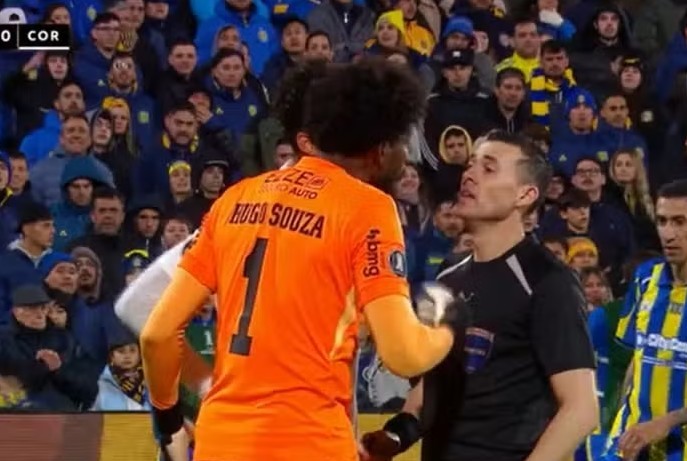 Goleiro Hugo Souza, de camisa laranja com número 1, discute com um árbitro de camisa preta em campo de futebol, com torcida ao fundo