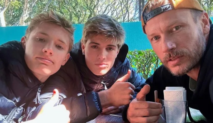 Três homens, dois jovens e um adulto, posam para uma selfie ao ar livre. O adulto, à direita, usa boné virado para trás e barba. Os três vestem jaquetas escuras e fazem sinal de positivo com as mãos. Ao fundo, vegetação e um muro azul