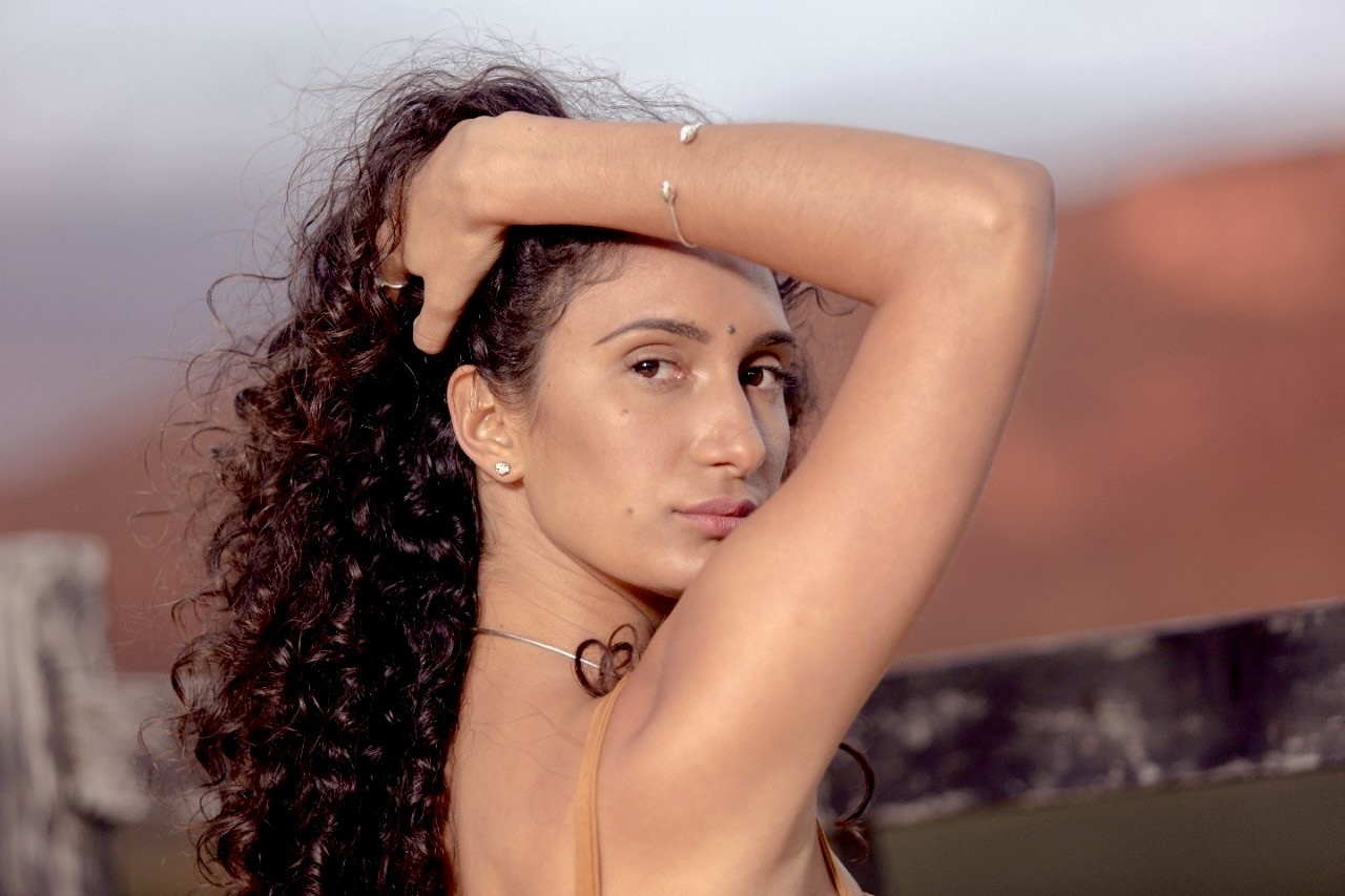 Mulher de pele clara, cabelos castanhos cacheados, com a mão direita na cabeça e o braço esquerdo levantado, olhando para a frente. Usa brinco de pérola e pulseira prateada com pérolas. Ao fundo, tons de marrom e cinza desfocados