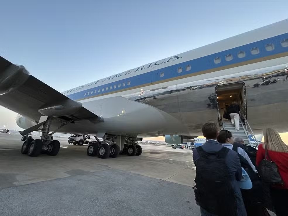 Pessoas embarcando em um avião grande, branco e azul, com a inscrição UNITED STATES OF AMERICA na lateral. A escada de embarque está aberta e uma pessoa sobe. Outras pessoas, de costas, aguardam na fila para entrar. O céu está claro e azul