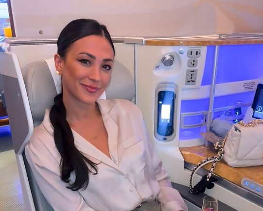 Mulher sorridente com cabelo escuro preso em rabo de cavalo, vestindo camisa de seda clara, sentada em assento de avião de primeira classe com iluminação azul e uma bolsa branca ao lado