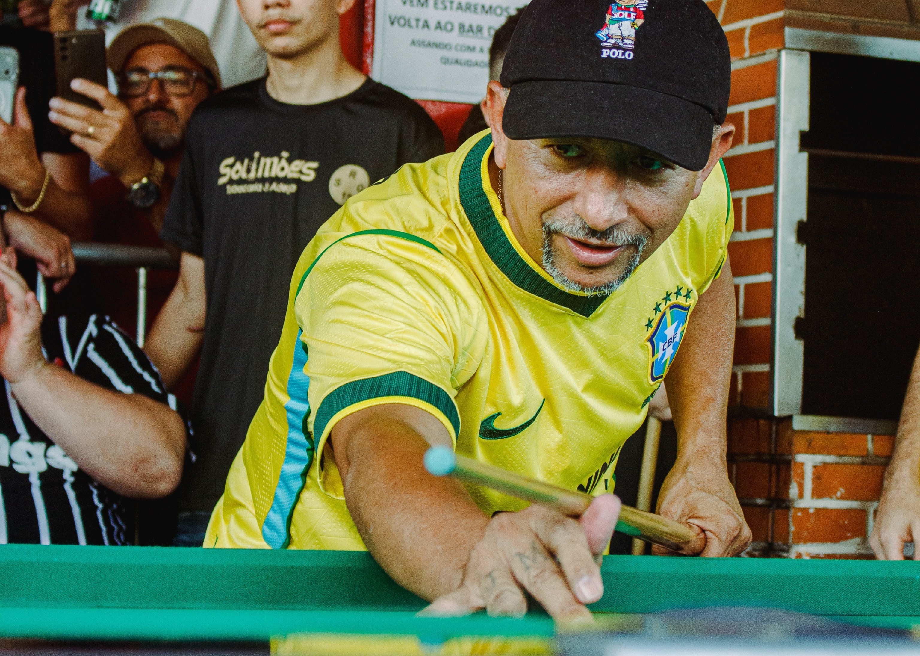 Homem de bon&eacute; preto e camisa amarela da sele&ccedil;&atilde;o brasileira joga sinuca, segurando o taco com a m&atilde;o direita sobre a mesa verde, enquanto olha para frente. Outras pessoas observam ao fundo
