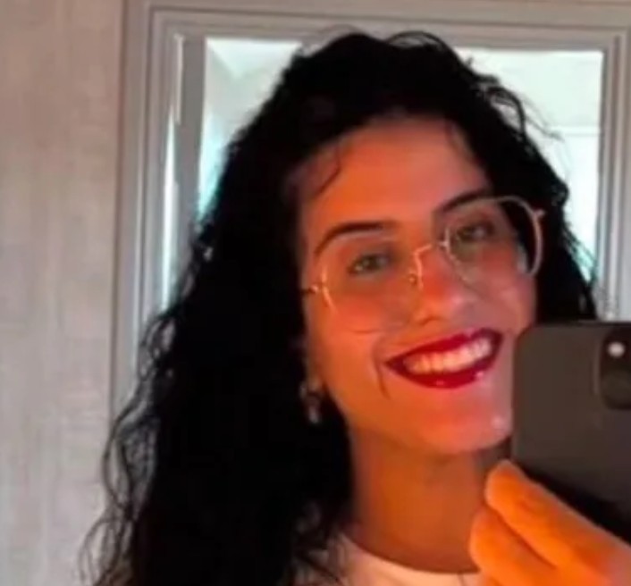 Mulher de cabelos escuros e cacheados, com óculos de armação dourada, sorrindo e segurando um celular preto para tirar uma selfie