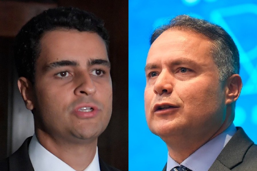 Dois homens em close-up, o da esquerda com cabelo escuro e terno preto, o da direita com cabelo grisalho e terno azul, ambos com expressões sérias e boca entreaberta, como se estivessem falando