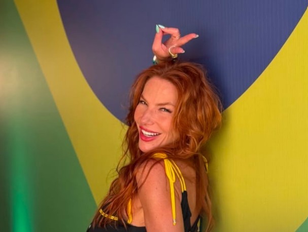 Mulher ruiva de cabelos longos, sorrindo e olhando para a câmera, com um top preto de alças amarelas. Ela levanta a mão direita acima da cabeça, fazendo um sinal de ok com os dedos, que estão com esmalte azul claro. Ao fundo, uma parede com as cores verde, amarelo e azul, remetendo à bandeira brasileira