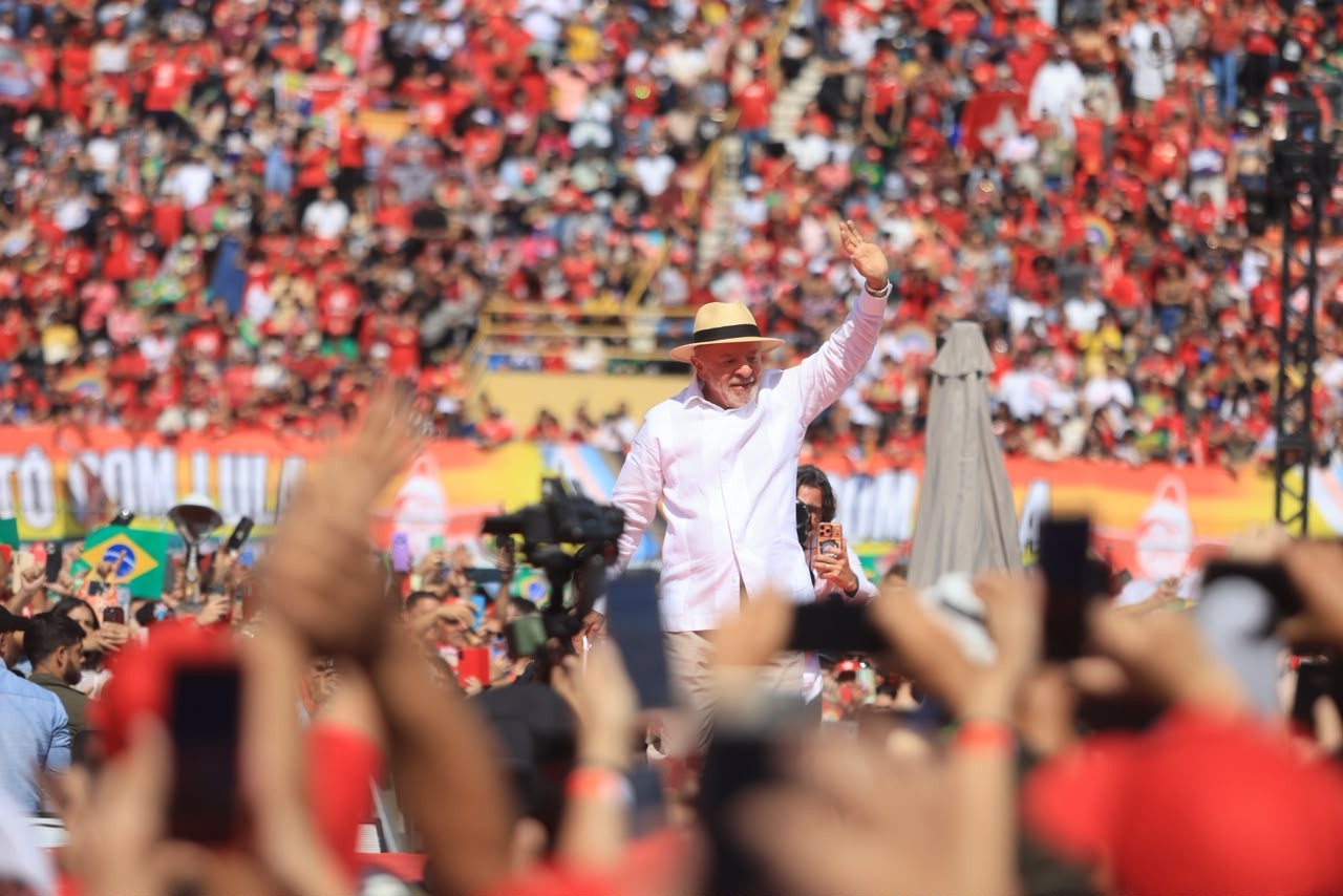 Lula, sorrindo e acenando com a mão direita, veste camisa branca e chapéu de palha, em um palco. Ao fundo, uma multidão de pessoas, muitas vestindo vermelho, e algumas bandeiras do Brasil. Mãos de pessoas na frente seguram celulares para filmar.
