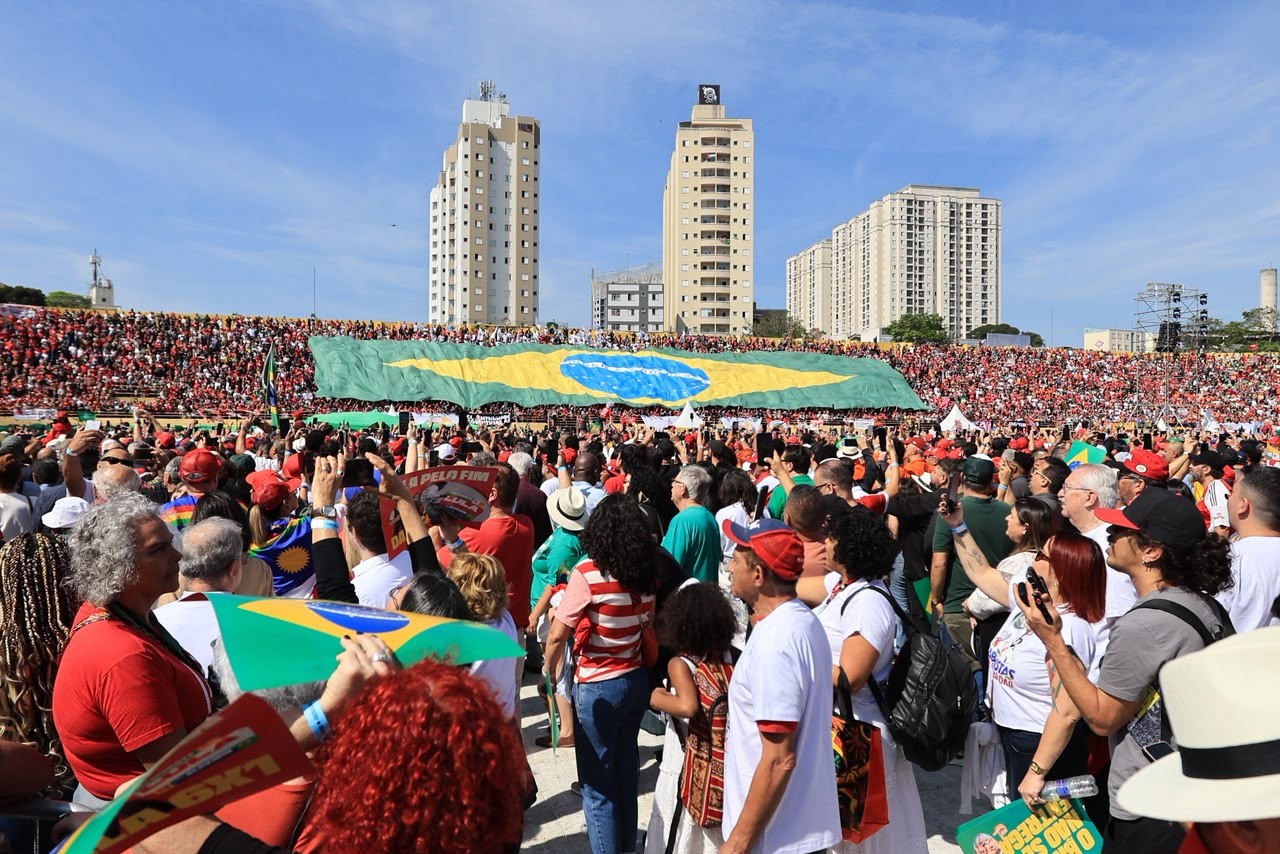 Multidão em um comício político, com muitas pessoas vestindo vermelho e segurando bandeiras do Brasil. Ao fundo, um grande estandarte verde e amarelo com o círculo azul da bandeira brasileira é exibido, e prédios altos se destacam sob um céu azul claro.