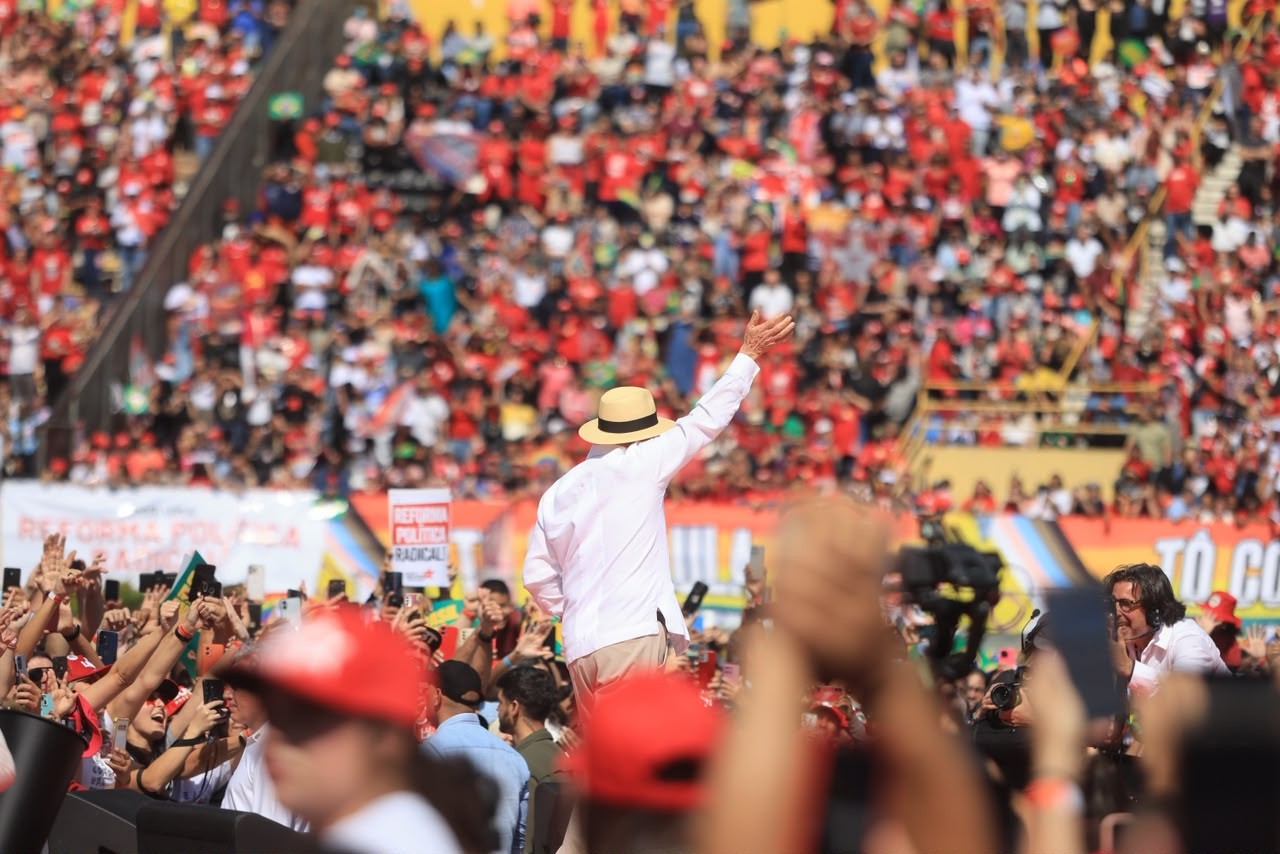 Homem de camisa branca e chapéu de palha, de costas, acena para uma multidão em um comício político. A plateia, majoritariamente vestida de vermelho, levanta os braços e celulares para filmar. Faixas com dizeres políticos são visíveis ao fundo