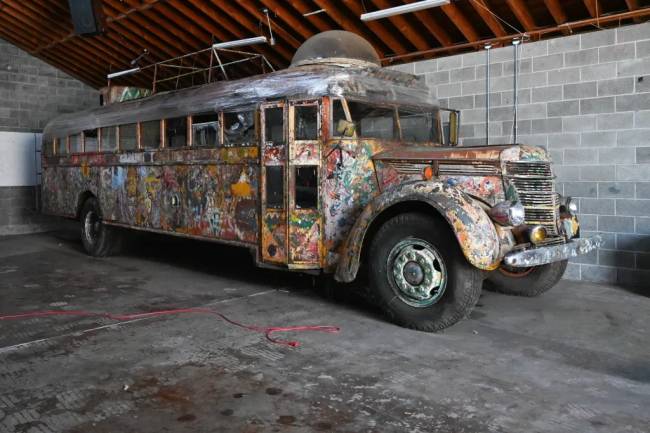&Ocirc;nibus antigo e enferrujado, coberto por grafites coloridos e uma c&uacute;pula transparente no teto, estacionado em uma garagem com paredes de blocos de concreto e teto de madeira