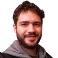 Homem branco de barba e cabelo castanhos, sorrindo levemente, vestindo um moletom cinza com capuz sobre uma jaqueta preta