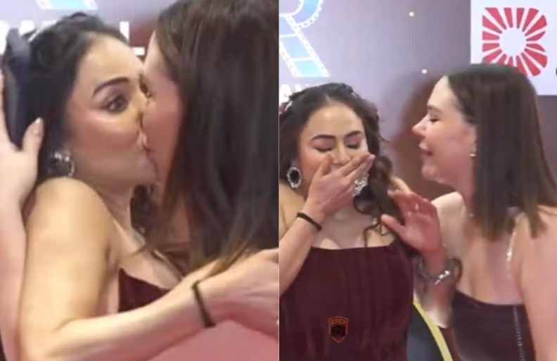 Duas mulheres em um evento. À esquerda, uma mulher de cabelo escuro e vestido vinho beija outra mulher. À direita, a mulher de vestido vinho cobre a boca com a mão, rindo, enquanto a outra mulher sorri ao seu lado
