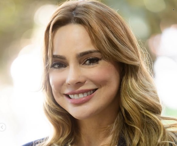 Mulher de cabelos castanhos claros e ondulados, sorrindo com os dentes à mostra, maquiagem leve e bochechas rosadas. Ela veste uma blusa escura e o fundo está desfocado com pontos de luz