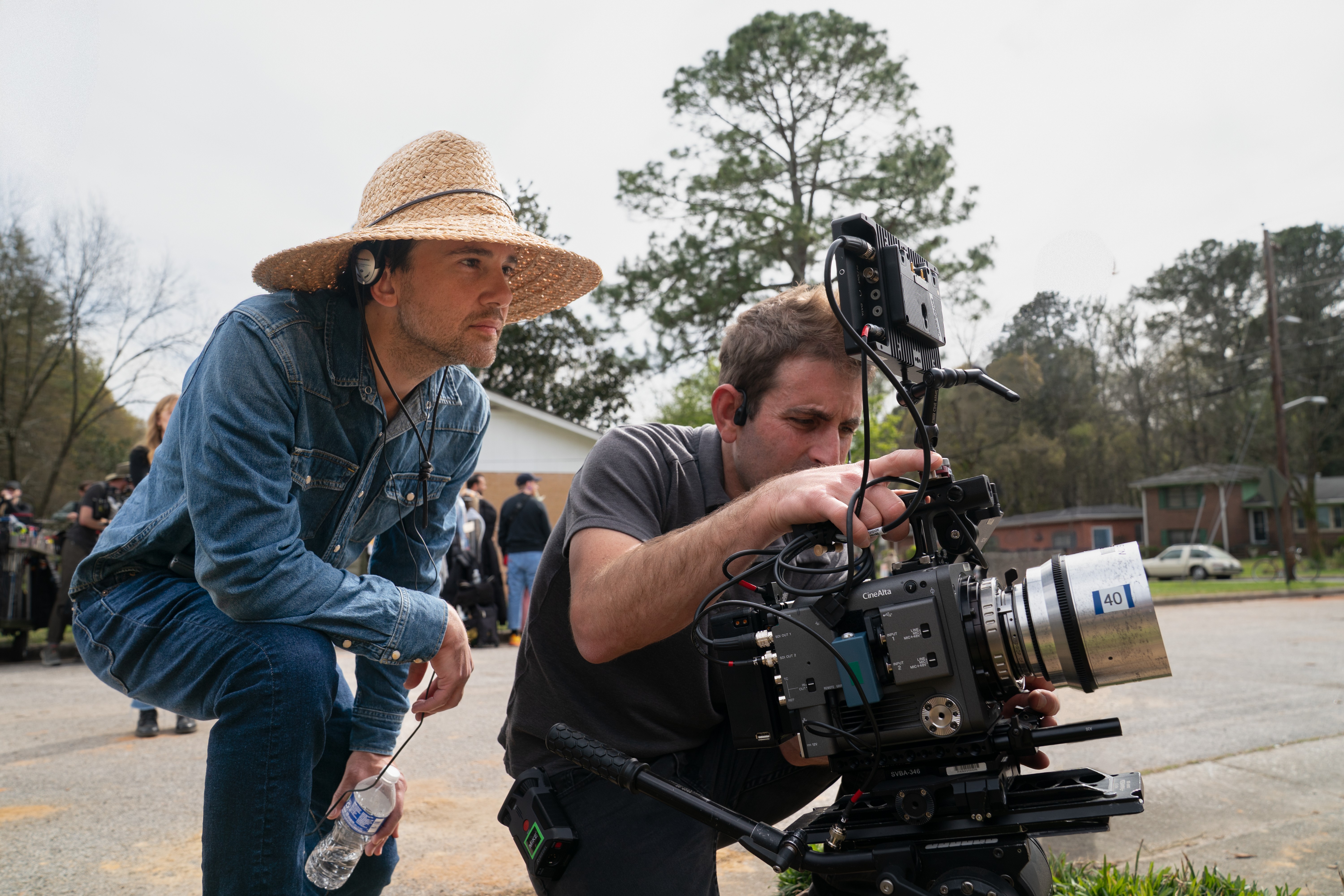 Dois homens trabalhando em uma filmagem externa. Um, de chapéu de palha e roupa jeans, segura uma garrafa de água. O outro, de camiseta cinza, ajusta uma câmera profissional em um tripé. Ao fundo, árvores e casas em um dia claro