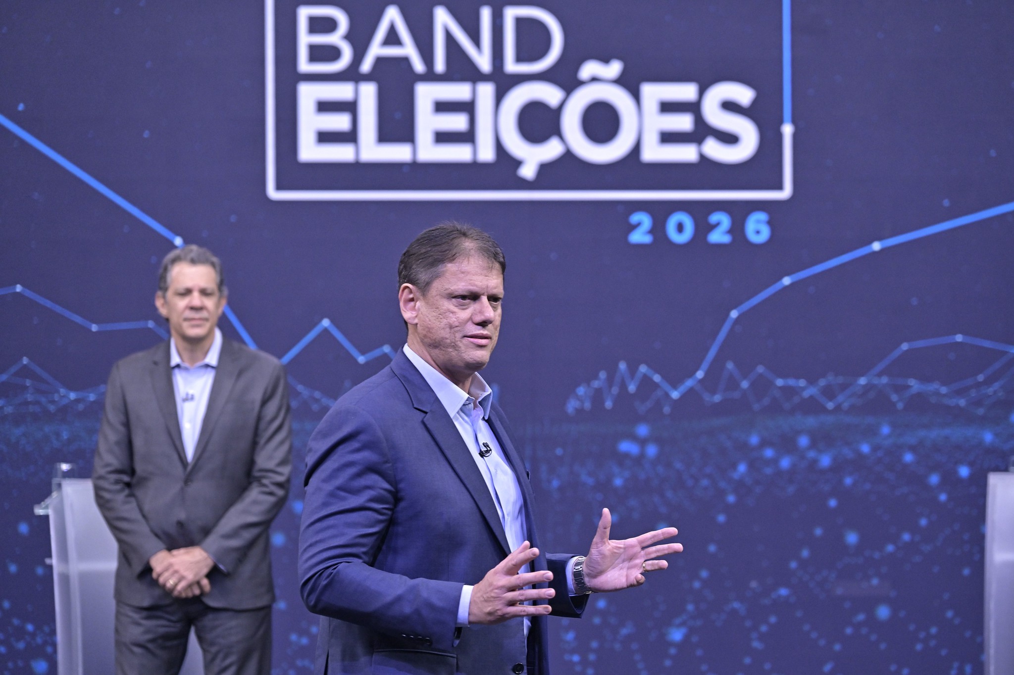 Dois homens em um palco azul escuro com gráficos e o texto BAND ELEIÇÕES 2026. Um homem de terno azul gesticula enquanto fala, e outro de terno cinza está parado atrás, com as mãos cruzadas