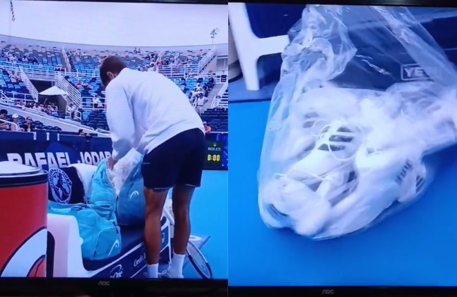 Um homem de costas, vestindo moletom branco e shorts escuros, se inclina para pegar algo em uma bolsa de tênis azul em uma quadra. À direita, um par de tênis brancos está dentro de um saco plástico transparente sobre uma superfície azul