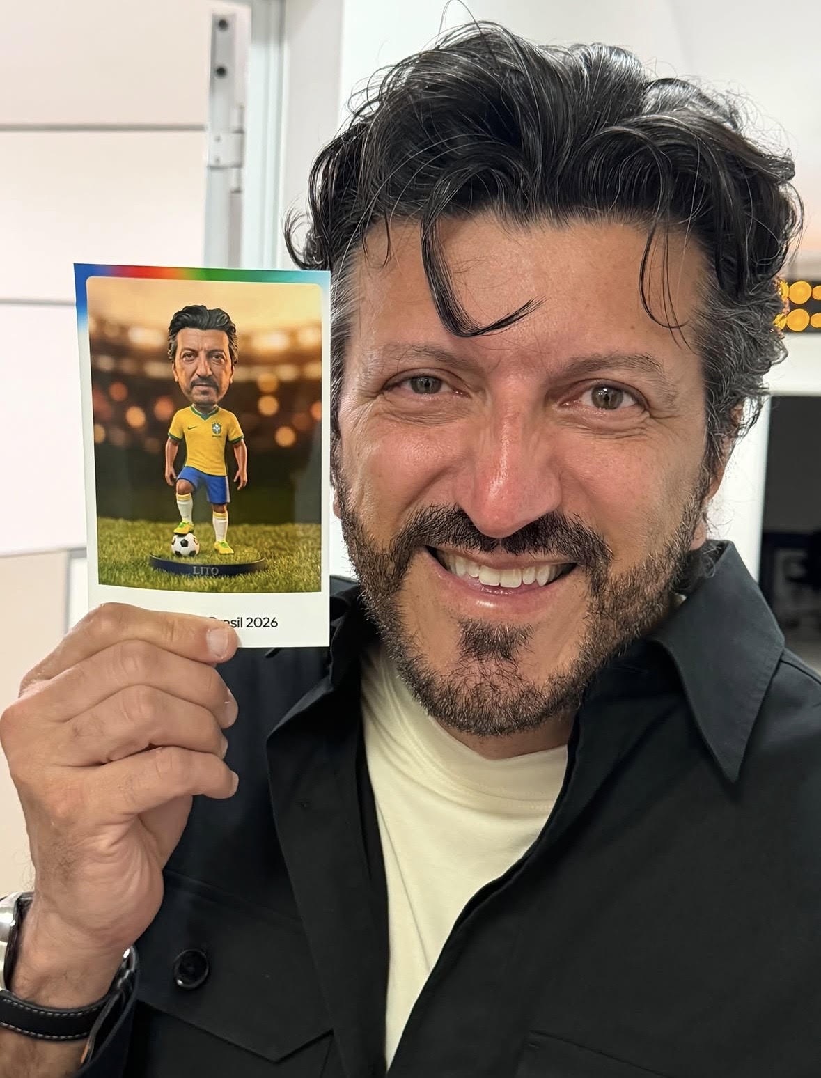 Homem de barba e cabelo escuro, sorrindo, segura um cartão com uma caricatura de si mesmo vestindo uniforme de futebol do Brasil em um campo