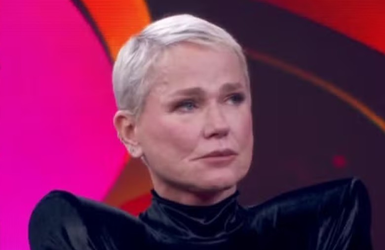 Xuxa Meneghel, mulher branca com cabelo curto platinado, veste blusa preta de gola alta e ombros marcados, olhando para o lado direito com express&atilde;o s&eacute;ria. O fundo &eacute; abstrato em tons de rosa e vermelho