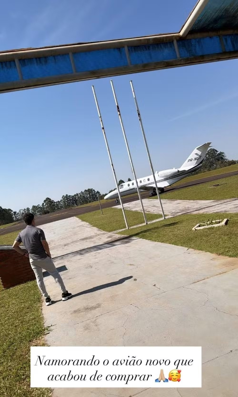 Homem de camisa cinza e calça clara de costas, observando um avião branco com a matrícula PR-NGT em uma pista de grama e asfalto, sob céu azul. Três mastros brancos e uma estrutura metálica com tela azul aparecem em primeiro plano. O texto sobreposto diz: Namorando o avião novo que acabou de comprar