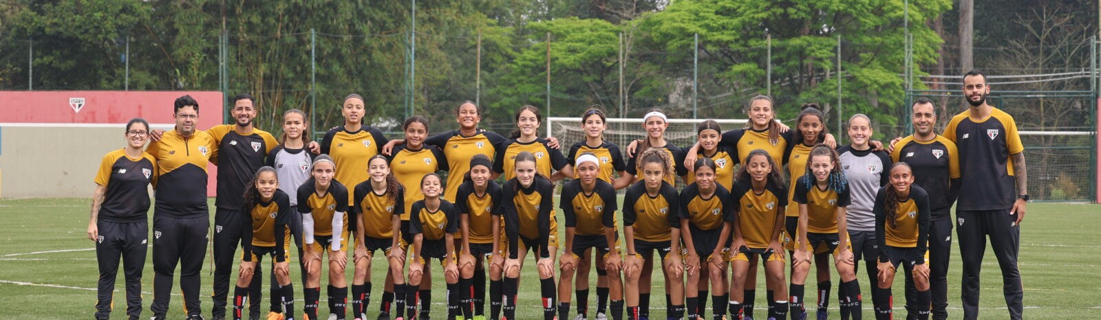 equipe de futebol feminino com cerca de 20 meninas e 5 adultos, todos sorrindo e usando uniformes pretos e amarelos, posando em campo gramado com árvores ao fundo