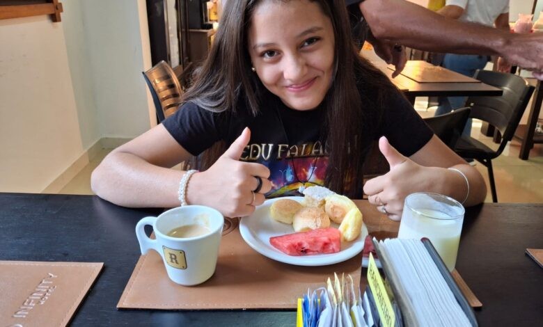 Jovem sorridente de cabelo castanho e camiseta preta, sentada à mesa, faz sinal de positivo com as duas mãos. À frente, um prato com pães e melancia, uma xícara de café e um copo de leite.