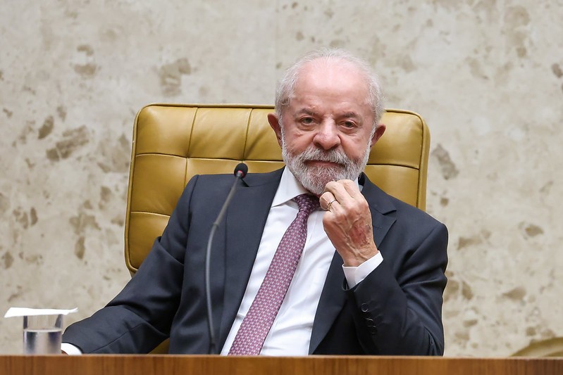 Luiz Inácio Lula da Silva, com barba e cabelos brancos, veste terno escuro e gravata roxa, sentado em uma cadeira de couro caramelo. Ele apoia a mão direita no queixo, com expressão pensativa, em um ambiente formal com parede de mármore claro e uma mesa de madeira à frente