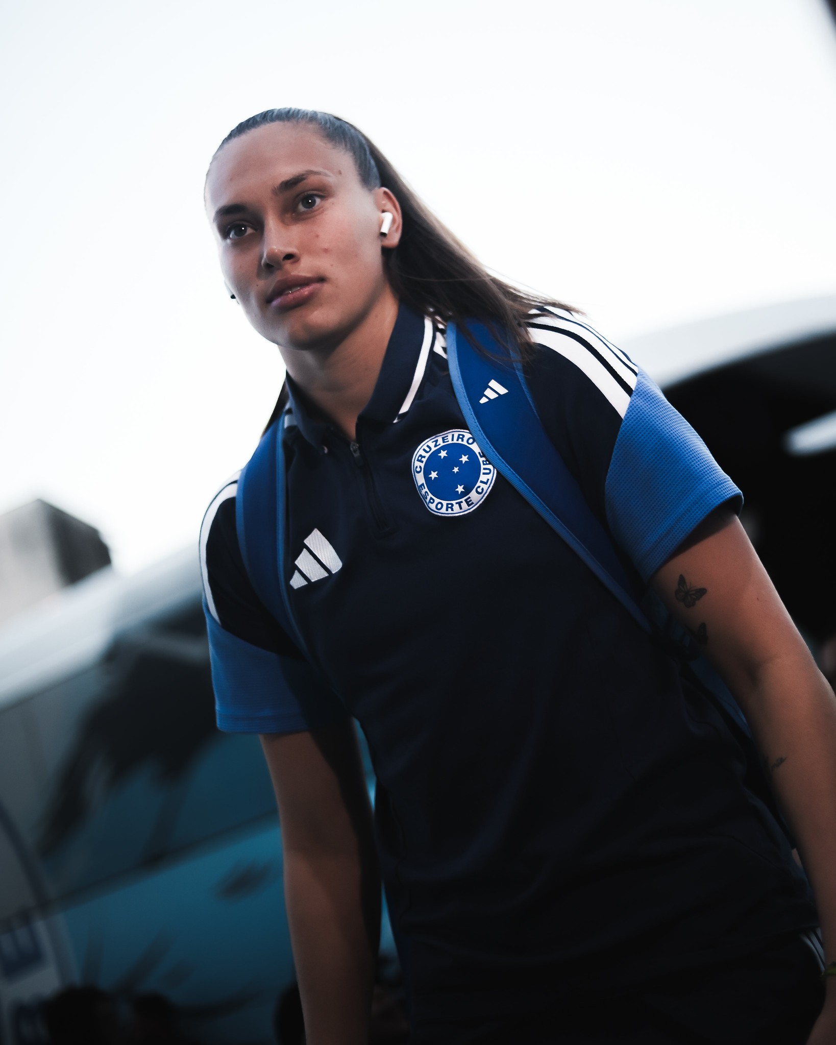 Mulher com rabo de cavalo, vestindo camisa polo azul-marinho do Cruzeiro e mochila azul, com fones de ouvido brancos, olhando para frente com express&atilde;o s&eacute;ria
