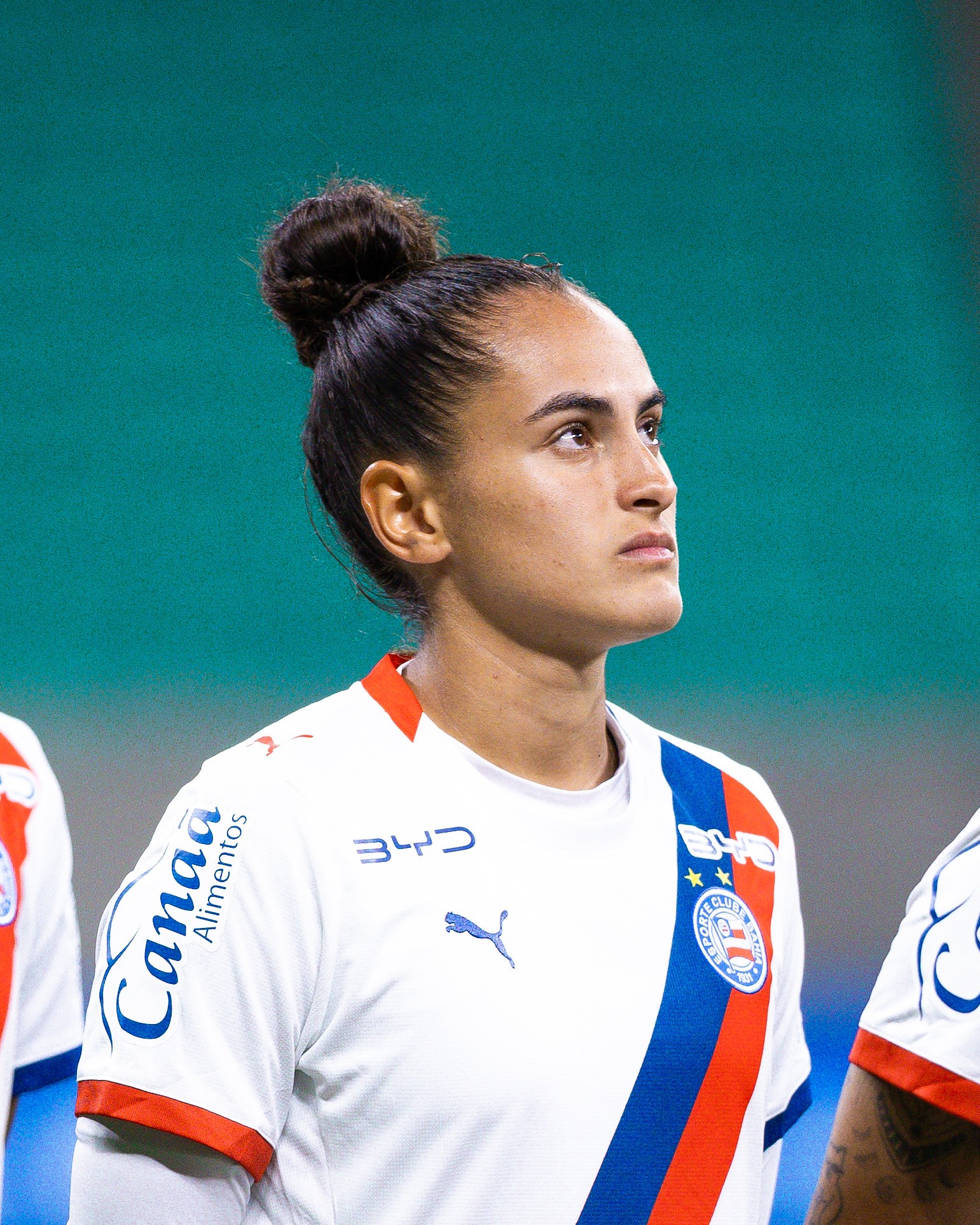 Jogadora de futebol com cabelo preso em coque, vestindo camisa branca do Bahia com listras azuis e vermelhas, olhando para cima com express&atilde;o s&eacute;ria em campo verde
