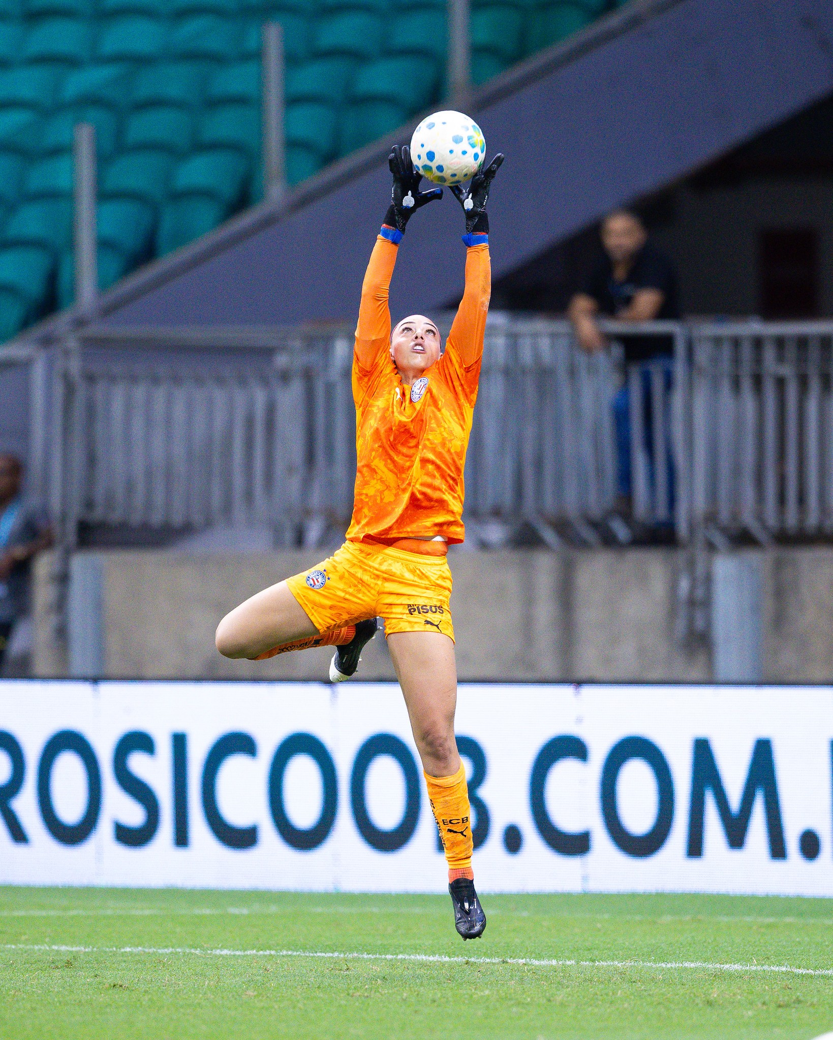Goleira de futebol feminino, vestindo uniforme laranja e luvas pretas, salta para agarrar uma bola branca com bolinhas azuis e amarelas. Ela est&aacute; no ar, com os bra&ccedil;os estendidos para cima, em um campo de grama verde com arquibancadas ao fundo