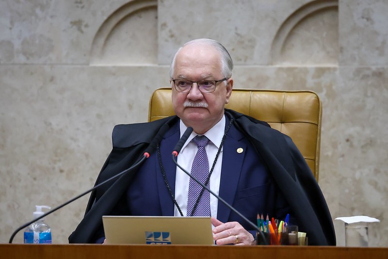 Homem idoso de óculos, bigode e cabelos brancos, vestindo terno azul, gravata roxa e toga preta, sentado em cadeira de couro marrom, com microfones à frente e um notebook sobre a mesa. Ele olha diretamente para a câmera com expressão séria