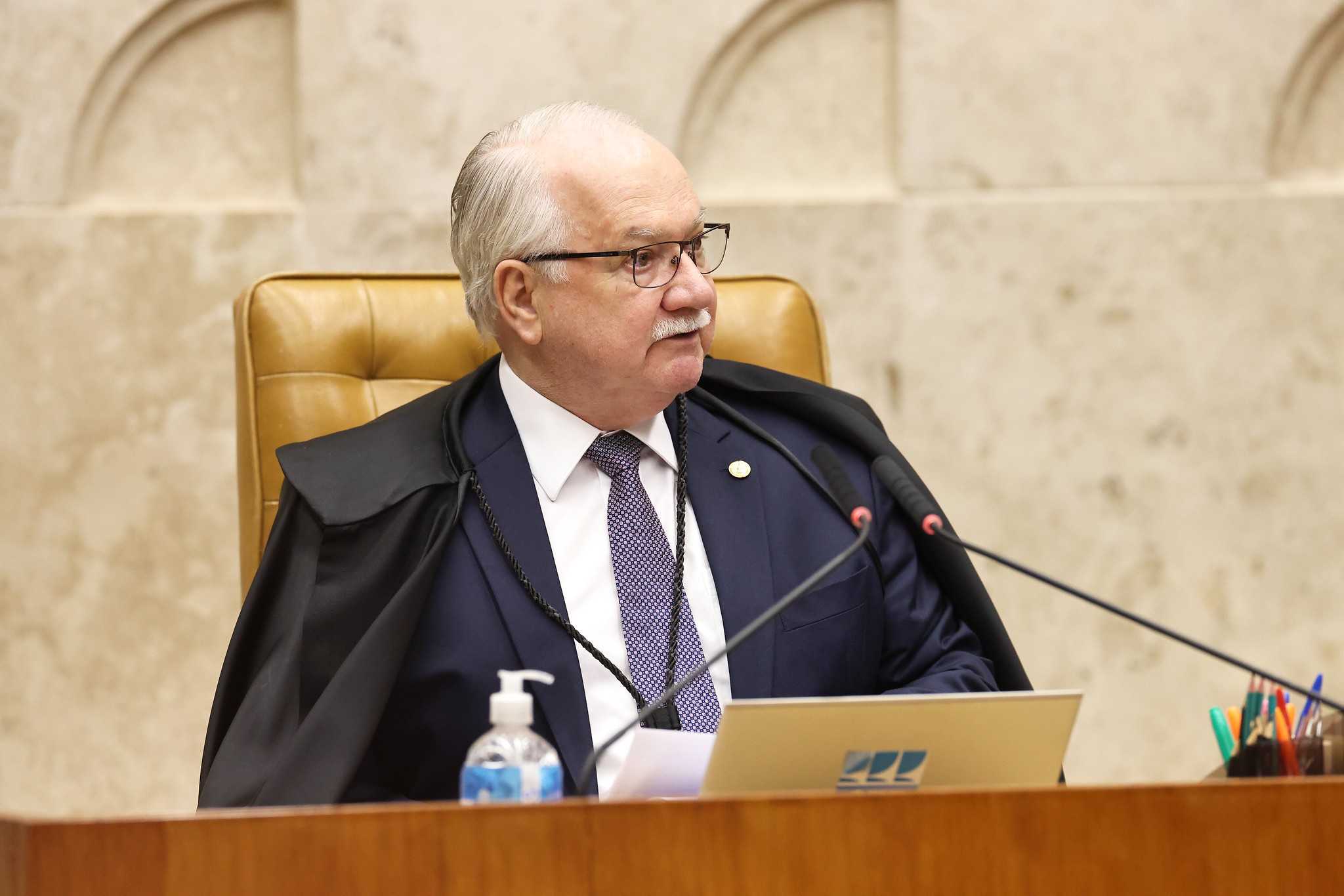 Homem idoso de cabelos brancos, bigode e óculos, vestido com terno azul-marinho, gravata estampada e toga preta, sentado em uma cadeira de couro marrom em um ambiente formal. Ele olha para a direita, com microfones à sua frente sobre uma mesa de madeira, ao lado de um frasco de álcool em gel e um porta-canetas