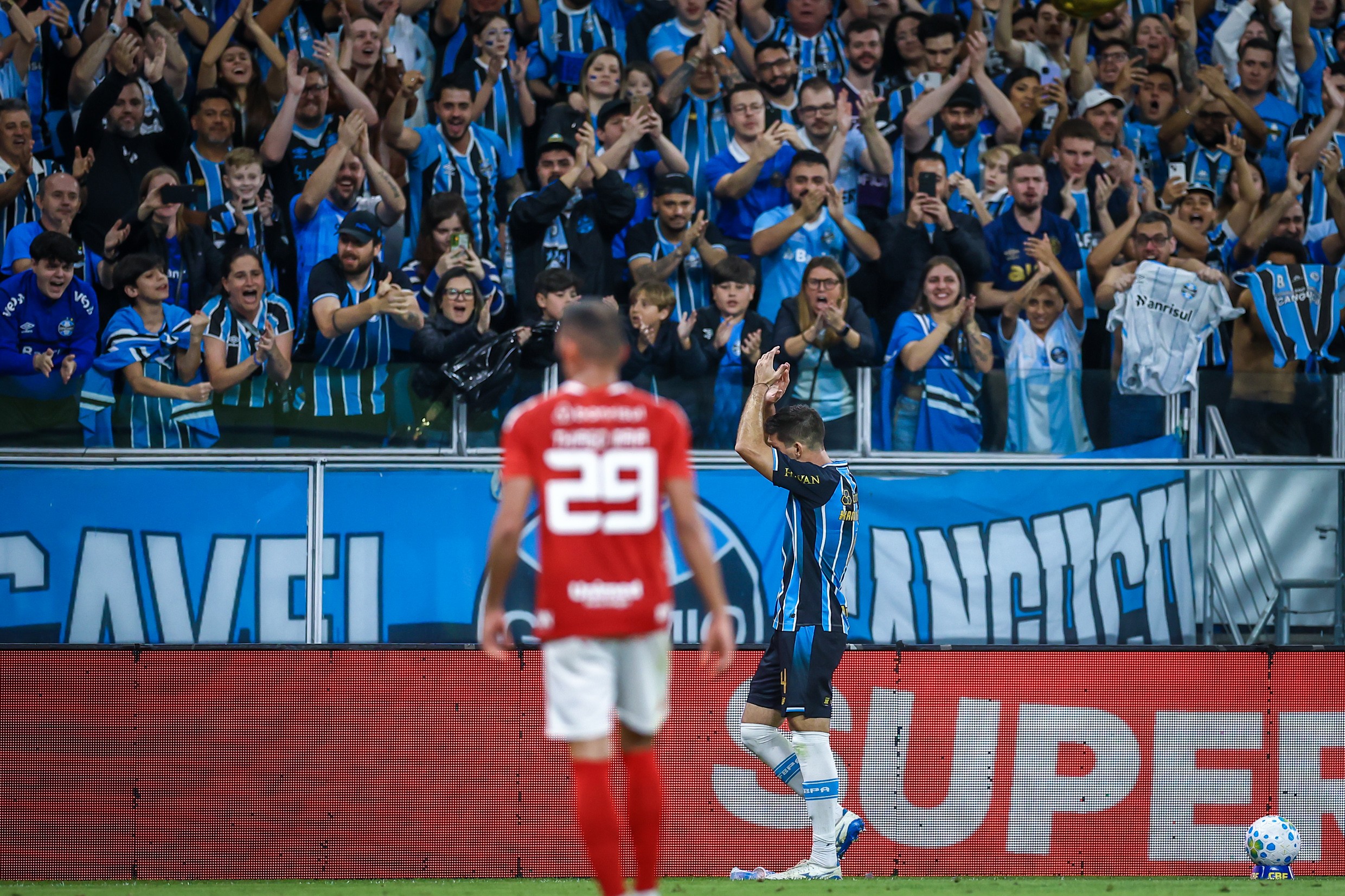 Torcida do Grêmio, vestindo camisas azuis e pretas, aplaude e vibra em um estádio de futebol. Em primeiro plano, dois jogadores, um de camisa vermelha com o número 29 e outro de camisa listrada azul e preta, caminham no campo. Uma bola de futebol está no canto inferior direito