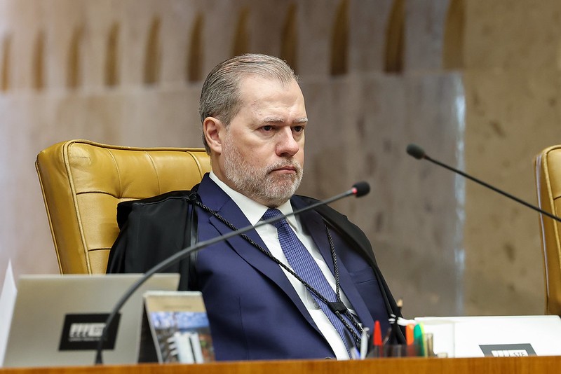 Um homem de meia-idade, com barba grisalha e terno azul, sentado em uma cadeira de couro marrom, com microfones à frente, em um ambiente formal de tribunal