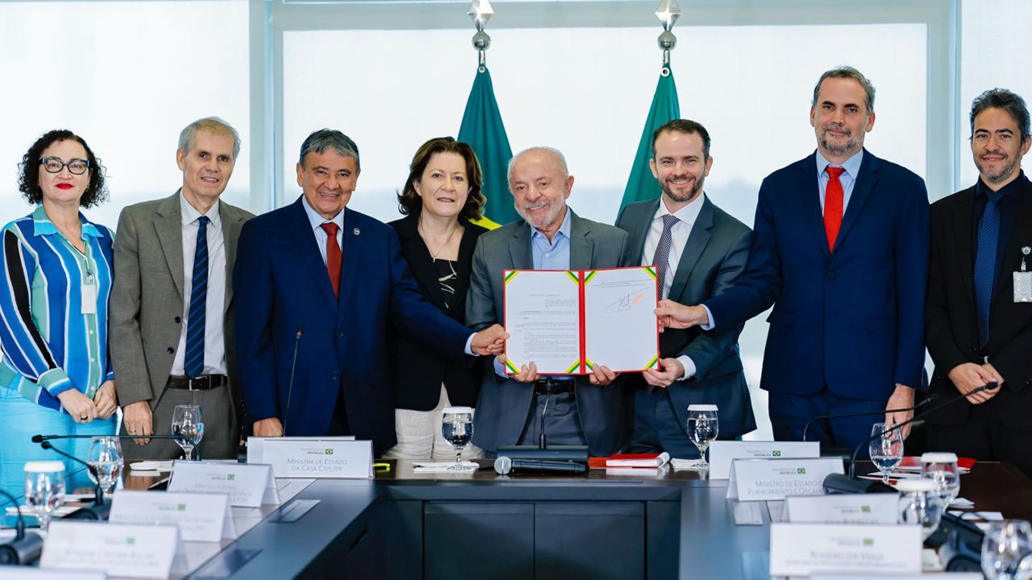 Nove pessoas, incluindo o presidente Lula, posam sorridentes atrás de uma mesa de reuniões, segurando um documento assinado. Duas bandeiras do Brasil estão ao fundo