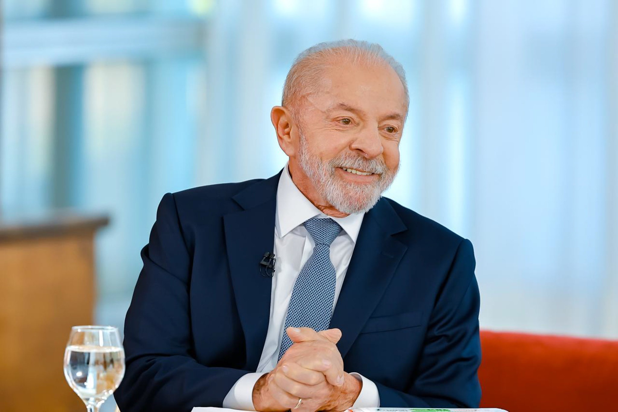 Homem idoso de barba branca, terno azul-marinho e gravata azul clara, sorrindo e com as mãos entrelaçadas sobre uma mesa, com um copo dágua à esquerda e fundo azul claro