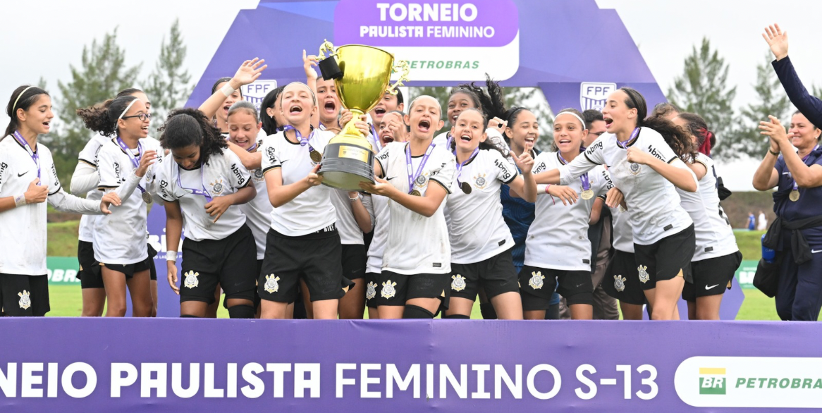 equipe de futebol feminino do Corinthians, com cerca de 15 jogadoras jovens e uma técnica, comemorando a triunfo no Torneio Paulista Feminino S-13. Elas vestem uniformes brancos e pretos, exibem medalhas no pescoço e erguem uma taça dourada, sorrindo e gritando de alegria em um pódio roxo com o logo da Petrobras