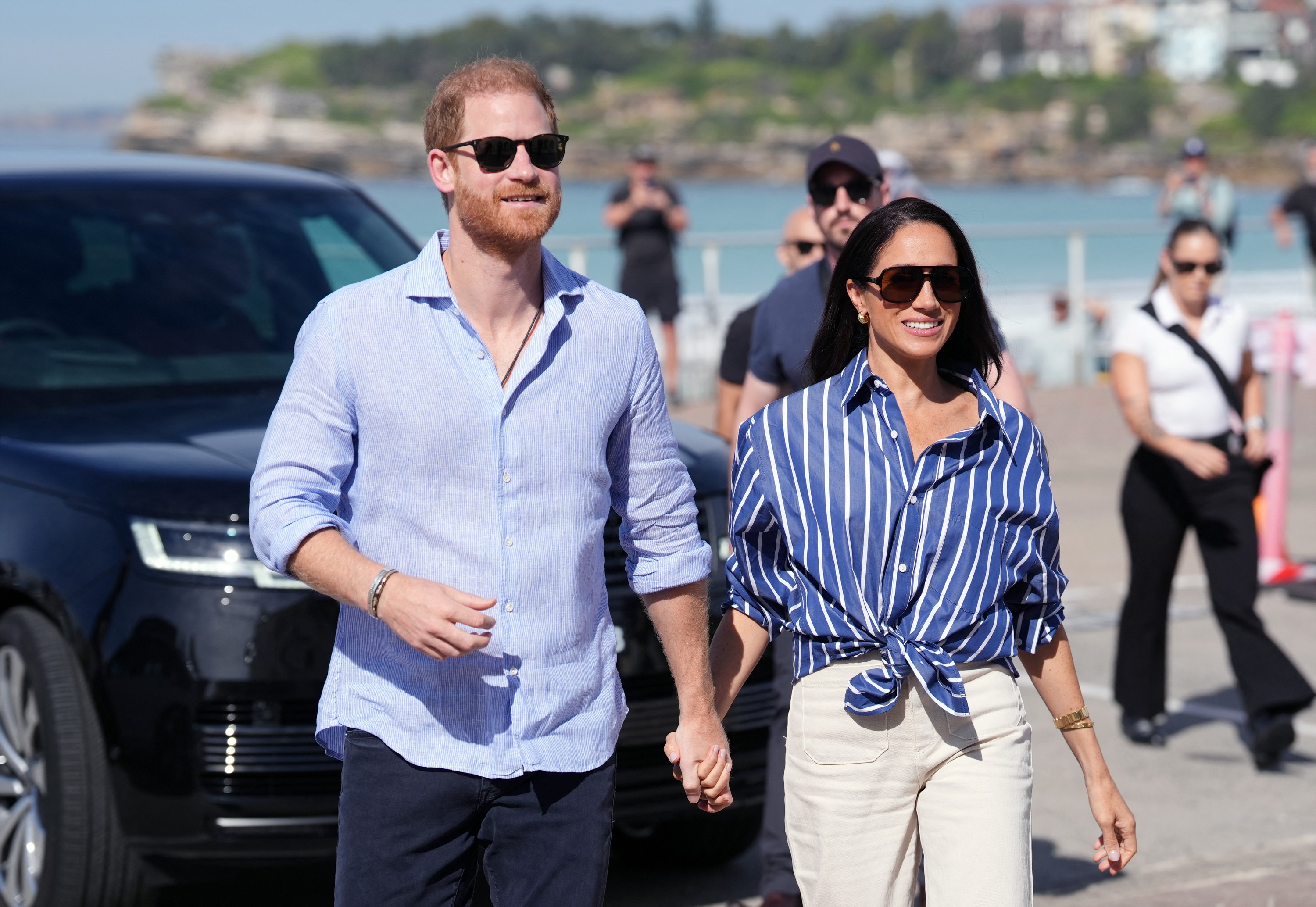 Príncipe Harry e Meghan Markle, sorridentes e de óculos escuros, caminham de mãos dadas em dia ensolarado. Harry veste camisa azul clara e calça escura. Meghan usa camisa azul listrada com nó na cintura e calça bege. Um carro preto está à esquerda e pessoas desfocadas ao fundo, com mar e prédios distantes