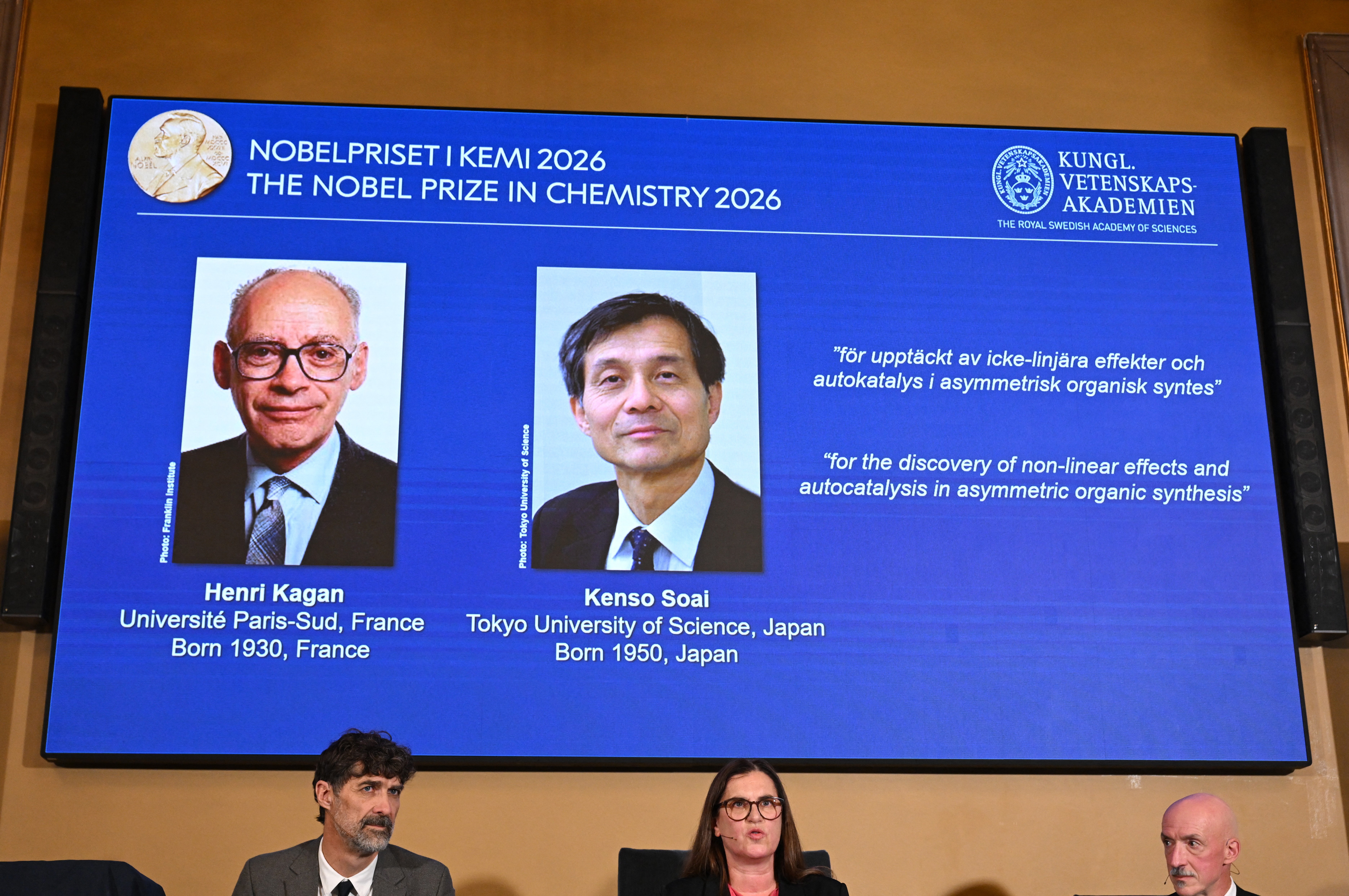 Tela azul exibe fotos de Henri Kagan e Kenso Soai, laureados com o Prêmio Nobel de Química 2026, por descobertas em efeitos não lineares e autocatálise. Abaixo, três pessoas assistem à apresentação