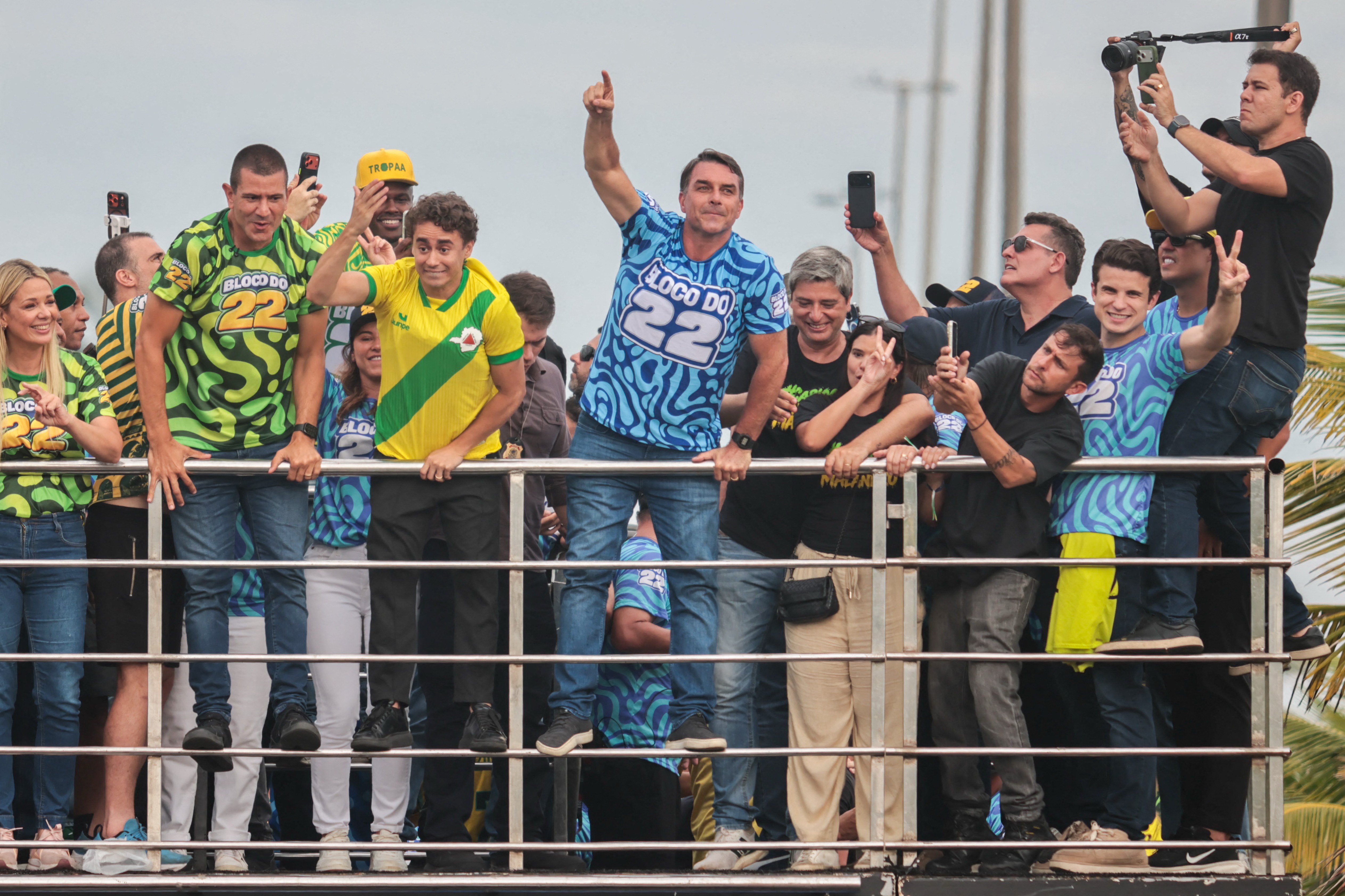 Grupo de pessoas em cima de um trio elétrico, com o ex-presidente Jair Bolsonaro no centro, vestindo camiseta azul com Bloco do 22. Ele aponta o dedo indicador para cima. Outras pessoas ao redor usam camisetas verdes e amarelas, algumas filmando com celulares ou câmeras.