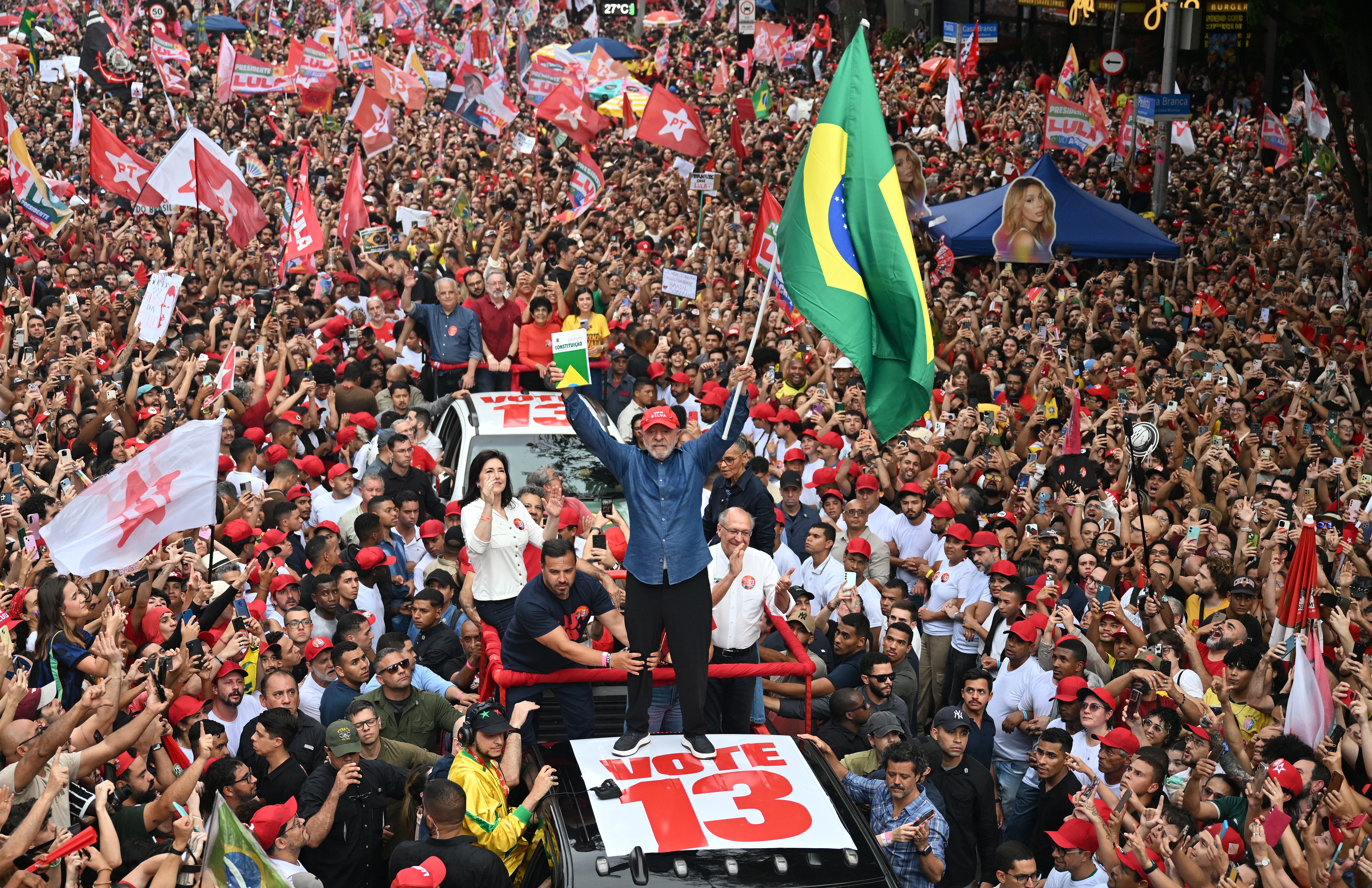 Lula, de camisa jeans e boné vermelho, acena com os braços erguidos sobre um carro com VOTE 13 em destaque, segurando uma bandeira do Brasil e um livro verde. Uma multidão de apoiadores, muitos com bonés vermelhos e bandeiras, o cerca em um comício ao ar livre.