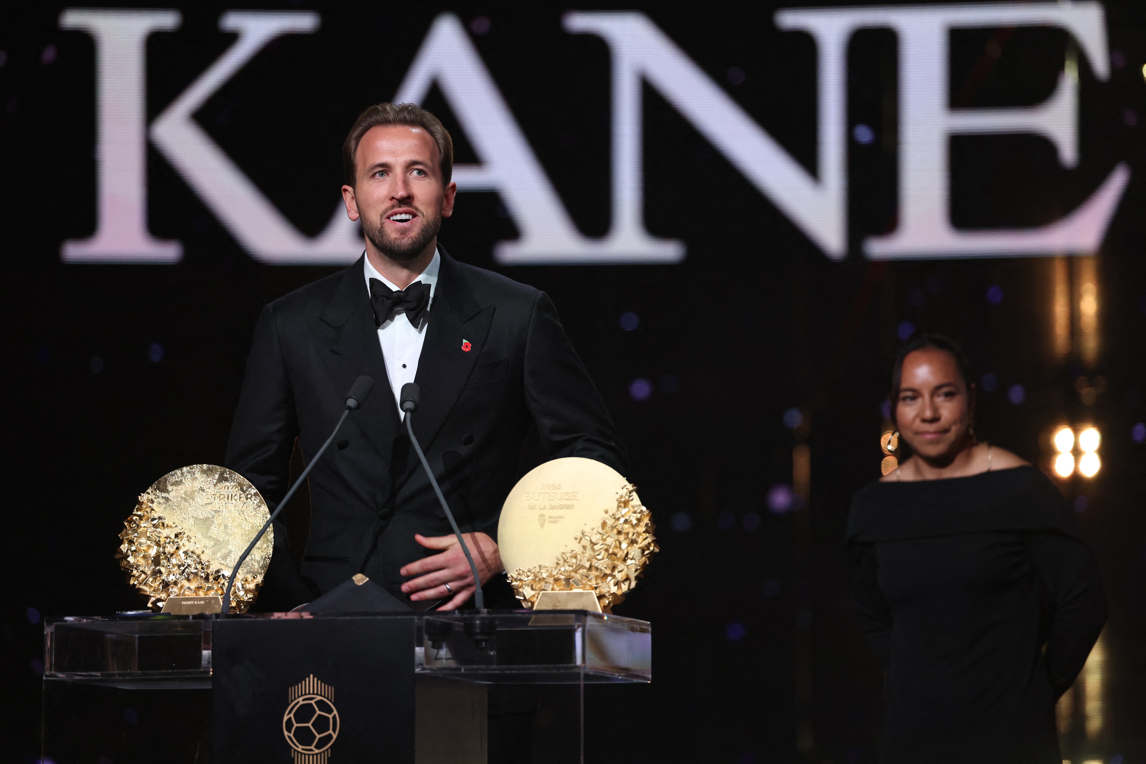 Harry Kane, de terno preto e gravata borboleta, fala ao microfone em um pódio com dois troféus dourados. Uma mulher de vestido preto e brincos grandes está à direita, sorrindo. O fundo escuro exibe a palavra KANE em letras brancas grandes