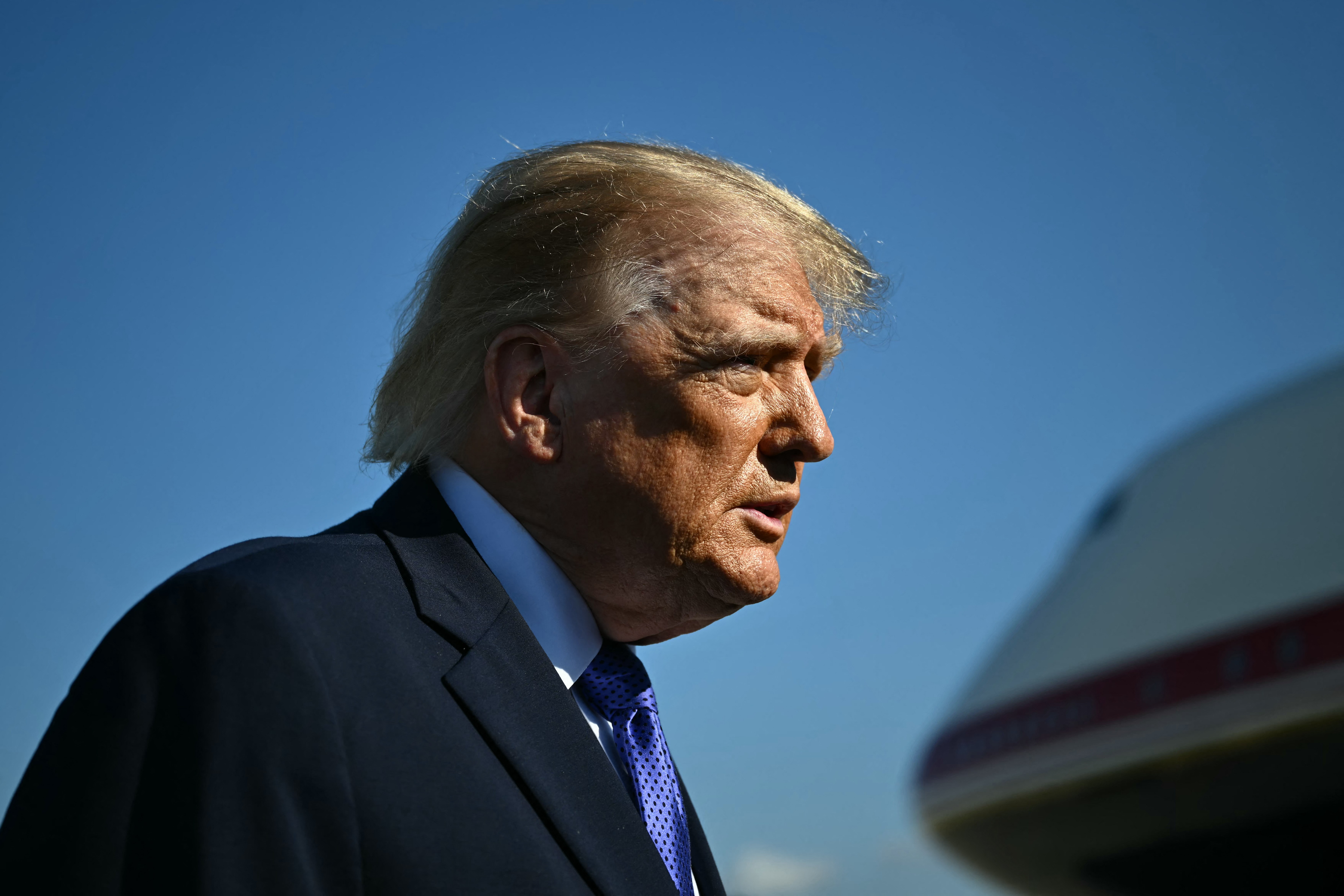 Donald Trump, com cabelo loiro e terno azul-marinho com gravata azul, é visto de perfil contra um céu azul claro, com parte de uma aeronave branca à direita