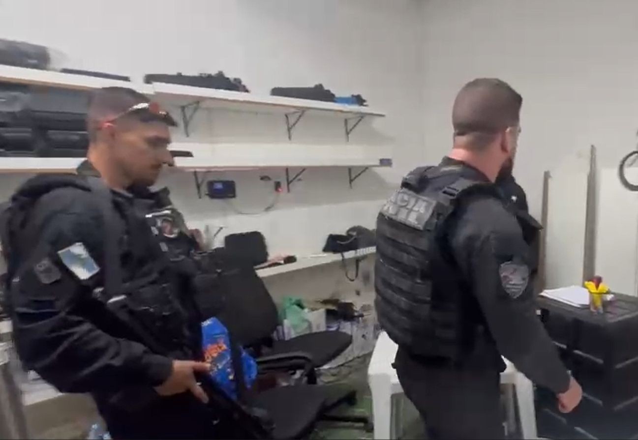 Dois policiais militares em uniforme preto e coletes balísticos, um de frente segurando um rifle e um pacote azul, e outro de costas, em um ambiente interno com prateleiras brancas cheias de objetos e uma mesa com papéis