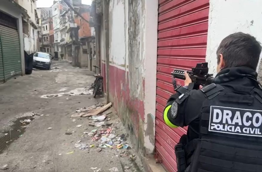 Policial da Polícia Civil, com colete tático preto e fuzil, mira em uma rua estreita e suja de uma comunidade, com prédios antigos e um carro branco ao fundo