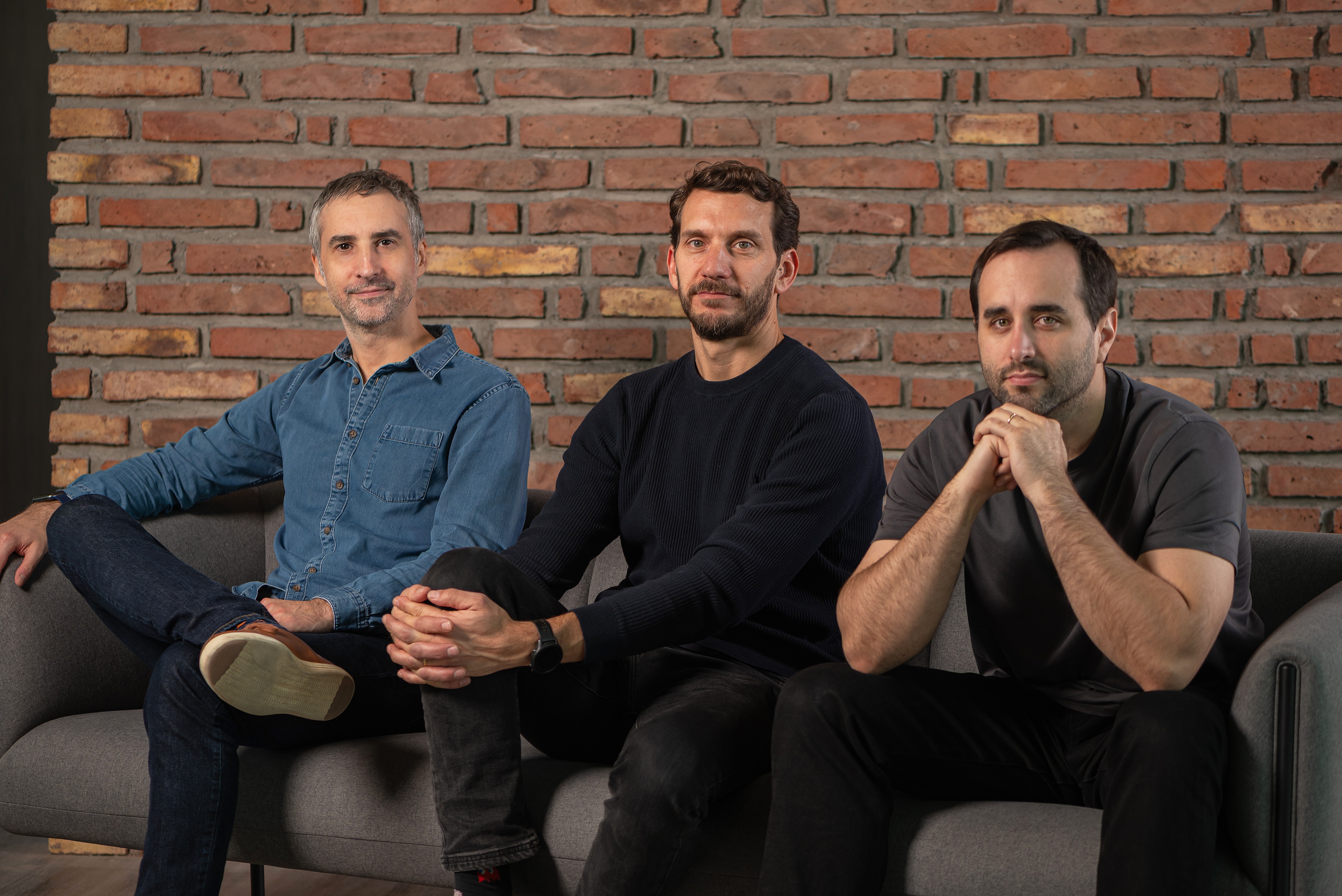 Três homens sentados em um sofá cinza, sorrindo para a câmera, com uma parede de tijolos à vista ao fundo. O da esquerda veste camisa jeans azul e calça escura, o do meio usa blusa de manga comprida preta e calça escura, e o da direita veste camiseta cinza escura e calça preta, com as mãos entrelaçadas sob o queixo