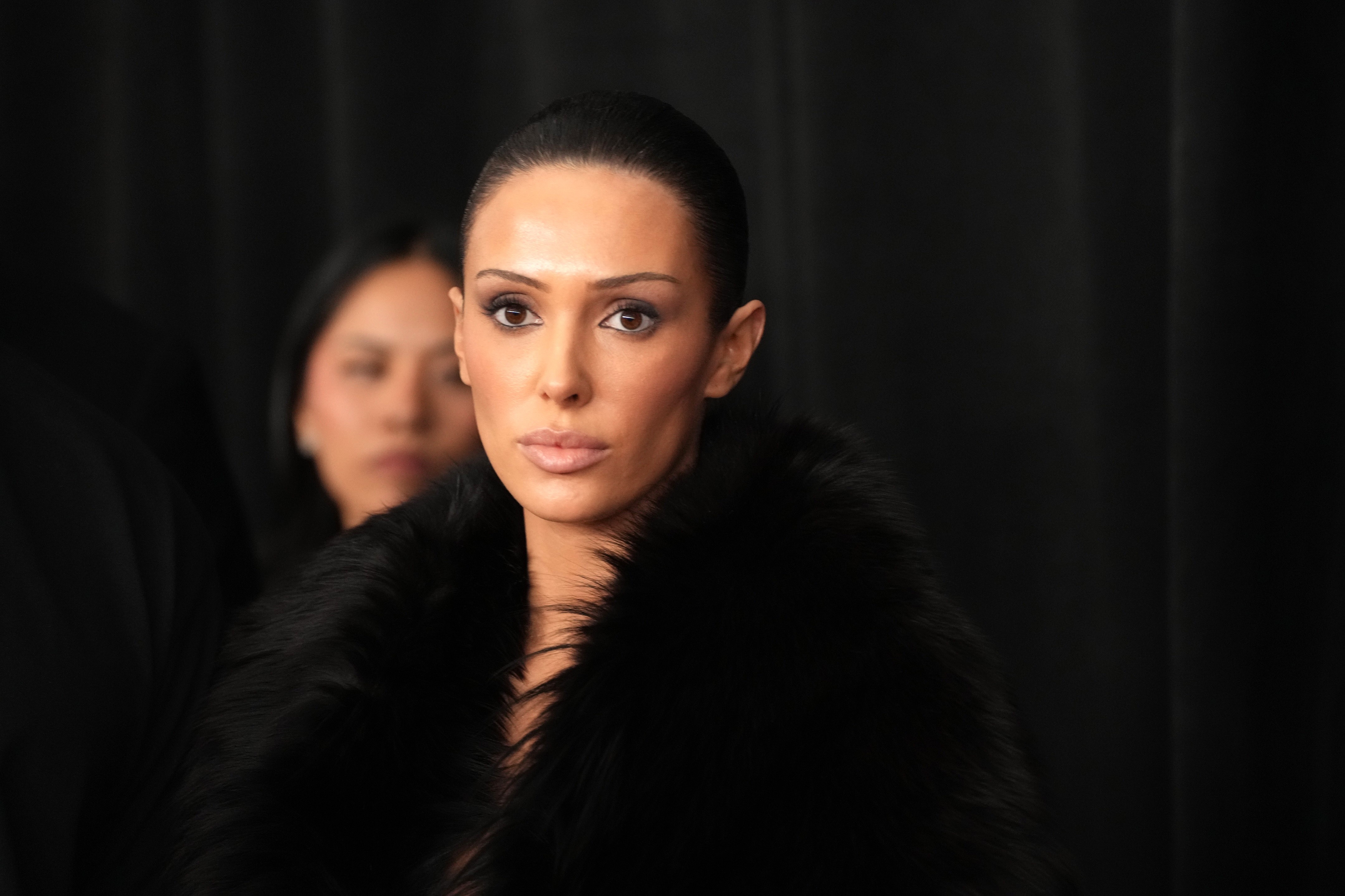 Mulher de pele clara e cabelo escuro preso, maquiagem marcante nos olhos, usando um casaco de pele preto, olhando diretamente para frente. Ao fundo, outra pessoa desfocada.