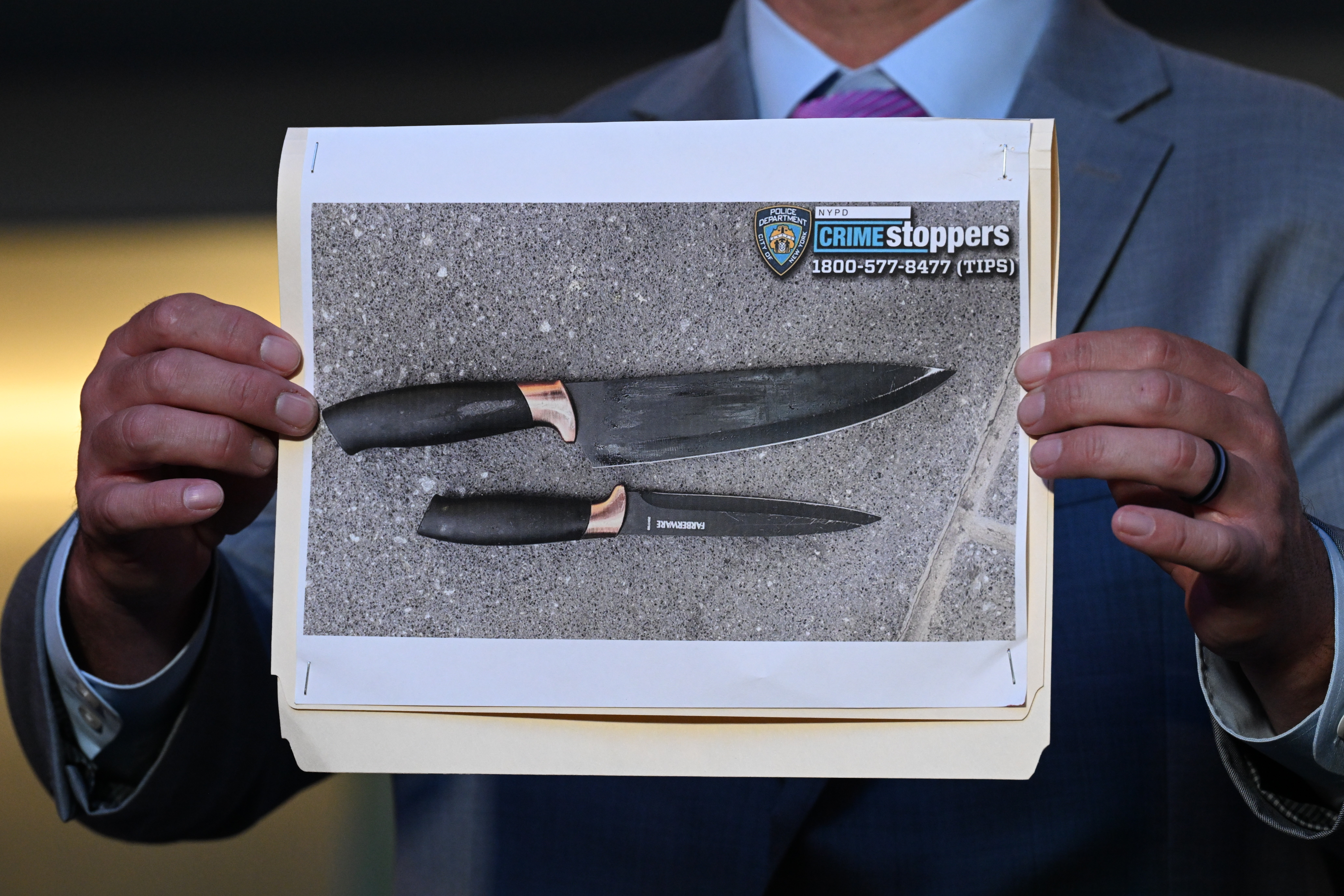 Homem de terno cinza e gravata roxa segura uma foto de duas facas de lâmina escura e cabo preto com detalhes em cobre, sobre um fundo cinza texturizado. No topo da foto, há um logo da NYPD e CRIME stoppers 1-800-577-8477 (TIPS)